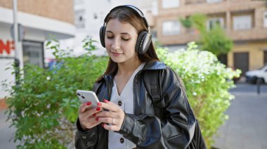 Genç bir kadın, şehir caddesinde akıllı telefon kullanırken kulaklıkla müzik dinlemekten zevk alıyor..