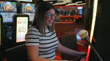 Gülümseyen genç bir İspanyol kadın canlı bir kumarhane ortamında kumar makinesiyle oynamaktan hoşlanıyor..