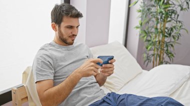 Sakallı, yakışıklı, yatak odasında akıllı telefon kullanan, rahatlama ve modern yaşam tarzını tasvir eden genç bir İspanyol..