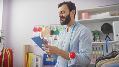 Sakallı orta yaşlı bir adam renkli bir terzi dükkanında tablet kullanıyor. İçi elbise ve dikiş aksesuarlarıyla dolu..