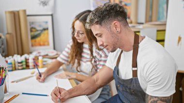 Bir adam ve bir kadın, renkli malzemelerle çevrili iyi donanımlı bir sanat stüdyosunda çizim yapıyorlar..