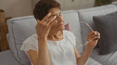 Kısa saçlı, yaşlı bir İspanyol kadın oturma odasında oturmuş gözlüğünü çıkartıyor. Evde sakin bir rahatlama anında yakalanmış..