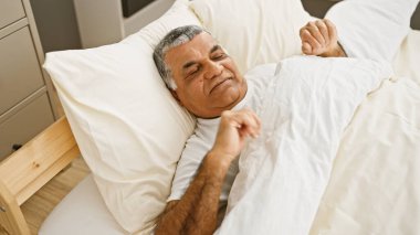 Olgun, gri saçlı bir adam, rahat, beyaz çarşaflı bir odada uyanıyor..
