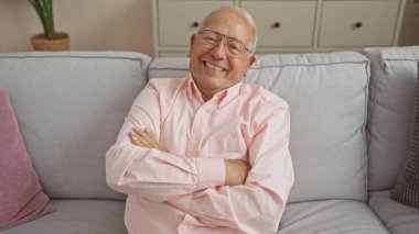 Beyaz saçlı, gözlüklü, orta yaşlı yakışıklı beyaz bir adam rahat bir oturma odasında kanepede otururken rahatça gülümsüyor..