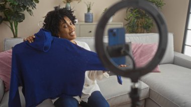 Güzel, kıvırcık saçlı Afro-Amerikan bir kadın oturma odasında video kaydederken mavi bir süveter tutuyor, rahat ev dekorasyonu ve ev bitkilerinin yer aldığı bir video..