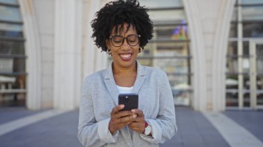 Kıvırcık saçlı, gözlüklü, açık bir şehir caddesinde akıllı telefonunu kullanırken gülümseyen güzel bir Afro-Amerikan kadını..
