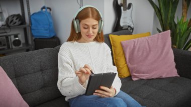 Genç kızıl saçlı kadın, kulaklık takıyor, modern evdeki rahat koltukta dijital tablet yiyor..
