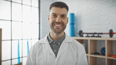 Beyaz önlüklü gülümseyen bir adam rehabilitasyon kliniğinde duruyor. Bu da profesyonel, sağlık hizmetleri ve terapi ortamını gösteriyor..