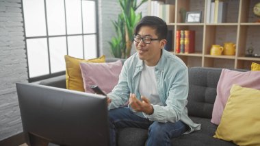 Genç Asyalı bir adam rahat, modern bir oturma odasında uzaktan kumandayla keyfine bakıyor.