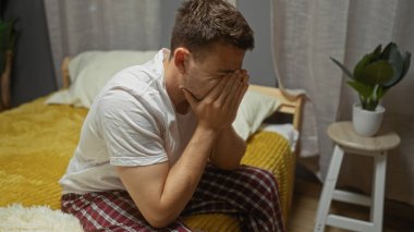 Sakallı genç bir İspanyol adam evdeki rahat bir yatak odasında yatağında oturuyor, elleriyle yüzünü örtüyor, beyaz bir gömlek ve kareli pantolon giyiyor, taburedeki küçük bir bitkinin yanında..