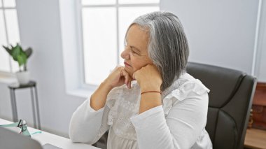 Serene kıdemli kadın modern ofis alanında elleri üzerinde dinleniyor, profesyonellik ve sakinlik yayıyor..