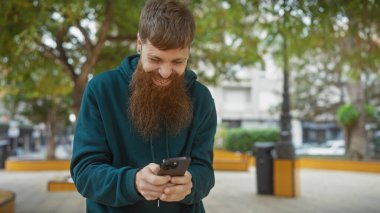 Sakallı kızıl saçlı bir adam yeşil şehir parkında akıllı telefonunu kullanırken gülümsüyor..