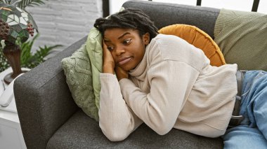 Düşünceli, genç, siyah bir kadın. Rastalı bir şekilde kanepede uzanıyor. Kapalı bir ortamda, huzur ve konfor hissi yayıyor..