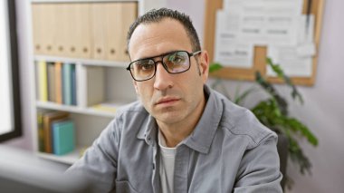 Gözlüklü ve gri gömlekli kendine güvenen İspanyol modern bir ofiste poz veriyor..