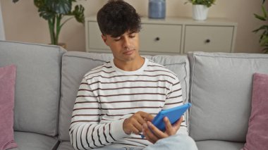 Oturma odasında oturan genç bir İspanyol adam, bitki ve dekorla çevrili rahat ve modern bir evle çevrili bir tablet kullanıyor..