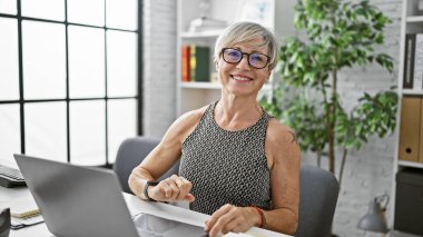 Gri kısa saçlı, gülümseyen, gözlüklü, kendine güvenen, ofis masasında oturan, profesyonel bir çalışma ortamını resmeden olgun bir kadın..
