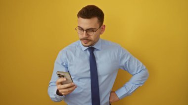 Sakallı, gözlüklü, mavi gömlekli ve kravatlı yakışıklı bir adam, izole edilmiş bir portrede canlı sarı bir arka planda akıllı telefonuna bakıyor..