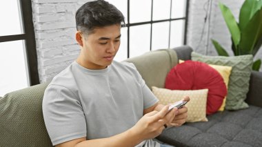 Odaklanmış Asyalı genç bir adam rahat bir şekilde akıllı telefonunu modern bir dairede kullanıyor..