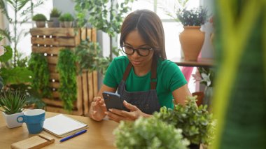 Yeşil bitkilerle dolu bir çiçekçide gözlüklü Afrikalı Amerikalı kadın akıllı telefon kullanıyor..