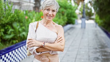 Kendine güveni tam, gri saçlı, yeşil bir parkta kollarını kavuşturmuş İspanyol bir kadın..