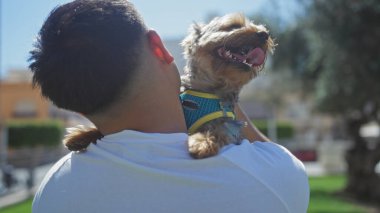 Genç Çinli bir adam şehir parkında güneşli bir günün tadını çıkarırken köpeğini yakın tutarak neşe ve bağlantı dolu hassas bir anı gösteriyor..