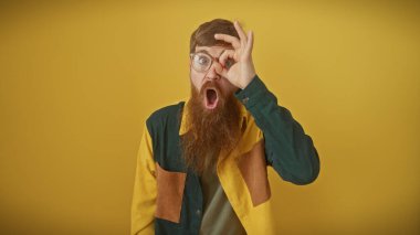 Gözlüklü, sakallı, yetişkin bir adamın sarı arka planda izole bir şekilde bir göze iyi bir jest yapmasına şaşırdım.