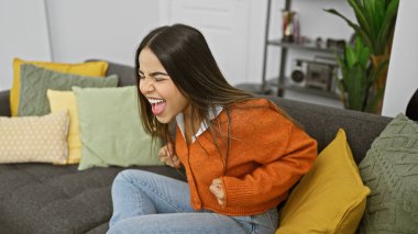 Neşeli, genç, İspanyol bir kadın rahat bir oturma odasında koltukta oturur ve coşkuyla güler..