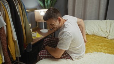 Yatak odasında sakallı, sırtı ağrıyan, kıyafetlerle çevrili ve komodinin üzerinde lamba yakan genç İspanyol bir erkek..