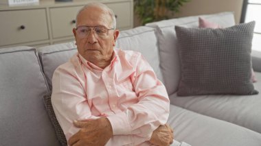 Olgun, gri saçlı, yaşlı, beyaz, düşünceli bir ifadeyle oturma odasında oturan, gözlüklü ve pembe gömlekli, ev dekorunu sergileyen bir adam..