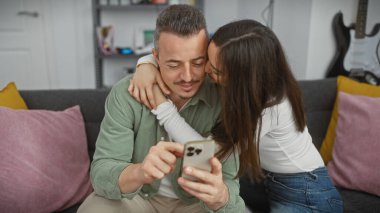 Sevgi dolu bir çift, rahat bir oturma odasında kucaklaşırken akıllı bir telefonla iletişim kurar. Bağlantı ve yakınlık uyandırır..