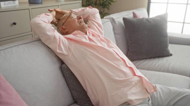 Rahat, gri saçlı, pembe tişörtlü yaşlı bir adam rahat bir oturma odasında rahat bir kanepede dinleniyor, elleri başının arkasında, evinin iç kısmının huzurunun tadını çıkarıyor..