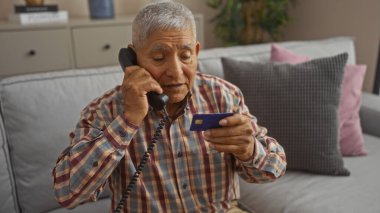 Oturma odasında elinde kredi kartıyla telefonla konuşan olgun İspanyol bir adam..