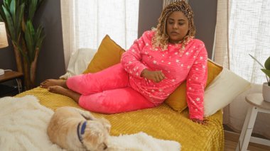 Pembe pijamalı Afro-Amerikalı genç bir kadın rahat bir yatak odasında köpeğiyle birlikte yatıyor..