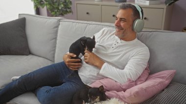 Orta yaşlı İspanyol bir adam oturma odasındaki kanepede dinlenirken iki chihuahua köpeği eşliğinde kulaklıkla müzik dinliyor..