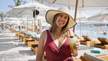 Şapkalı ve mayo giymiş gülümseyen bir kadın lüks bir havuz kenarında bir plaj kulübünde mojito tutuyor..