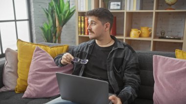 Bıyıklı ve gözlüklü, düşünceli bir İspanyol adam rahat bir oturma odasında elinde dizüstü bilgisayarla bir koltukta oturuyor..