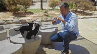 Orta yaşlı İspanyol bir adam akıllı telefonuyla diz çöker, şehir parkında iki chihuahuanın fotoğrafını çeker, evcil hayvanlarla sahipleri arasında samimi bir anı yakalar..