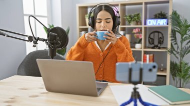 Latin bir kadın modern bir radyo stüdyosunda podcast atmosferini sembolize ederken kahvesini yudumluyor..