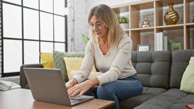 Kafkasyalı, olgun, sarı saçlı bir kadın rahat, modern bir oturma odasında dizüstü bilgisayarla çalışıyor.