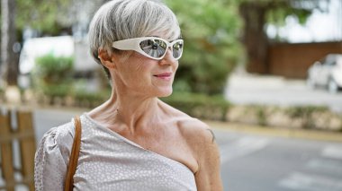 Kısa gri saçlı, güneş gözlüğü takan kendinden emin orta yaşlı bir kadın şehir ortamında poz veriyor..