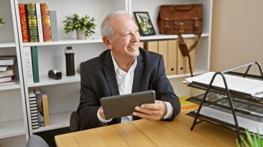 Kendinden emin ve neşeli kıdemli bir adam, beyaz saçlı akılda kalıcı bir patron, zarif ofis masasında dokunmatik dokunmatik pedini kullanarak mutlu bir şekilde çalışıyor, her gülüşünde başarı yayıyor..