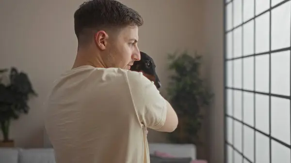 stock image A young hispanic man holding a dachshund dog in a cozy living room with large windows, indoor plants, and comfortable furniture.