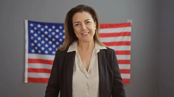 stock image A confident woman in a business suit stands proudly in an office with an american flag backdrop, embodying professionalism and patriotism.