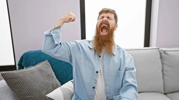 stock image Cheerful young redhead man confidently flexing strong biceps, radiating joy with a cool smile, while comfortably sitting in his homely living room.