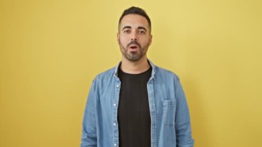 Genç bir İspanyol 'un komik portresi, yanakları şişiyor ve deli gibi surat yapıyor, şişmiş ağızla hava üflüyor. Kot gömlek giyiyor, sarı arka planda izole edilmiş..