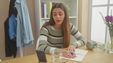 Genç İspanyol bir kadın, evde bir video görüşmesine katılmış, güneşli bir odada, odaklanmış bir ifadeyle zarifçe notlar yazıyor..