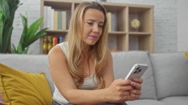 Rahat bir oturma odasında akıllı telefon kullanan düşünceli sarışın bir kadın..