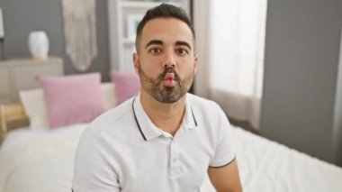 Komik genç İspanyol adam mutlu bir şekilde aptal bir balık suratı yapıyor, gözlerini kısıyor ve dudaklarını büzüyor, yatak odasında oturmuş, evde pozitif, çılgın bir titreşim yayıyor..
