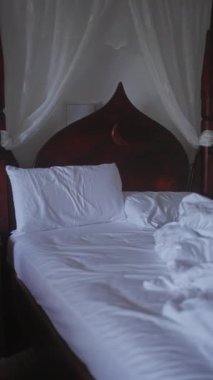 Yatak odasında beyaz çarşaflı, ahşap bir yatak başlığı ve doğal ışık ve katıksız perdelerle yapılmamış bir yatak..