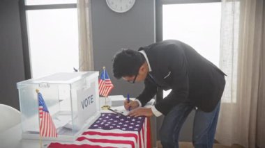 Takım elbiseli Asyalı genç bir adam Amerikan bayraklı bir Amerikan üniversitesi oy kullanma odasında oy kullanıyor..